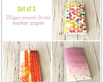 May include: Set of three 210gsm smooth Bristol marker paper pads. One pad has a geometric pattern with orange, yellow, red, and blue triangles. Another pad has a chevron pattern with orange, red, and white stripes. The third pad has a pink and purple ombre background with gold glitter dots.