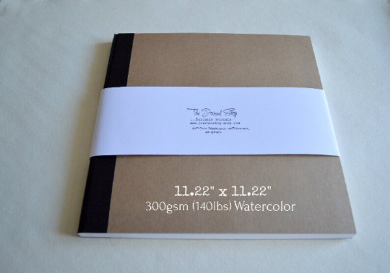 Puede incluir: Un cuaderno de bocetos de acuarela marr&oacute;n con bordes negros. La portada tiene el texto "The Paper Stop" y "11.22" x 11.22" 300 g/m&sup2; (140 lb) Acuarela" impreso en &eacute;l.