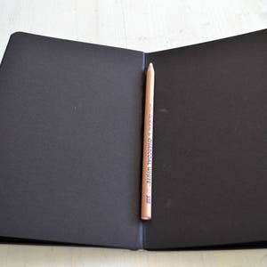 Pode incluir: Um caderno preto de capa dura com um l&aacute;pis saindo da lombada. O l&aacute;pis est&aacute; etiquetado como "General's Charcoal White 2B".