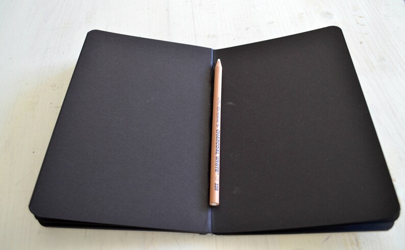 Pode incluir: Um caderno preto de capa dura com um l&aacute;pis saindo da lombada. O l&aacute;pis est&aacute; etiquetado como "General's Charcoal White 2B".