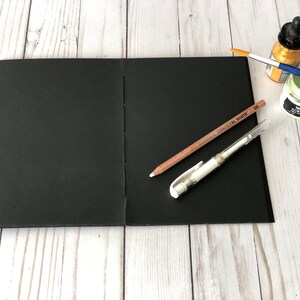May include: A black sketchbook with a white pencil, a white pen, a gold paint bottle, and a green paint jar on a white wooden surface.