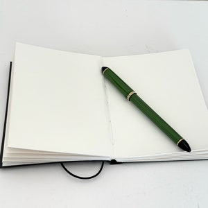 May include: A blank notebook with a black cover and a green pen lying open on a white surface.