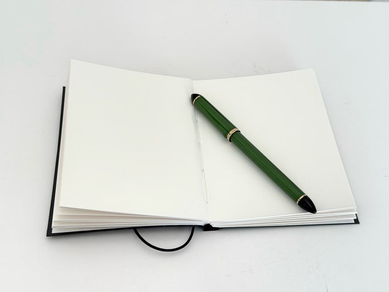 May include: A blank notebook with a black cover and a green pen lying open on a white surface.