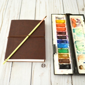 May include: A brown leather-bound journal with a yellow wooden pencil and a watercolour paint set with a variety of colours.