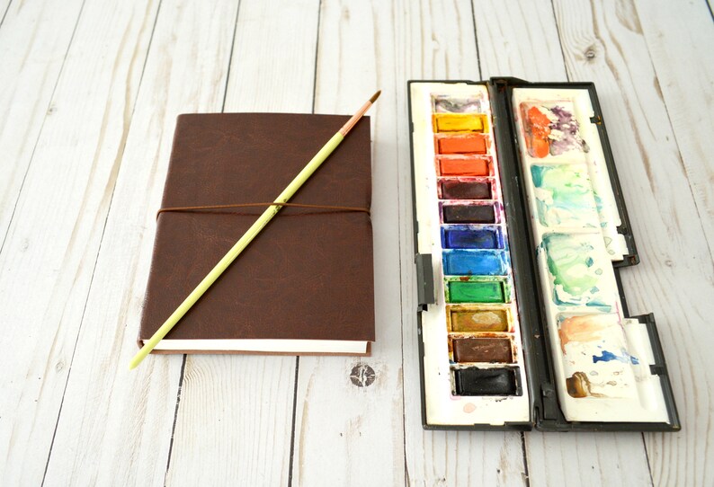 May include: A brown leather-bound journal with a yellow wooden pencil and a watercolour paint set with a variety of colours.