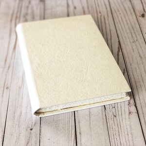 May include: A closed, off-white journal with a textured cover sits on a light wood surface.