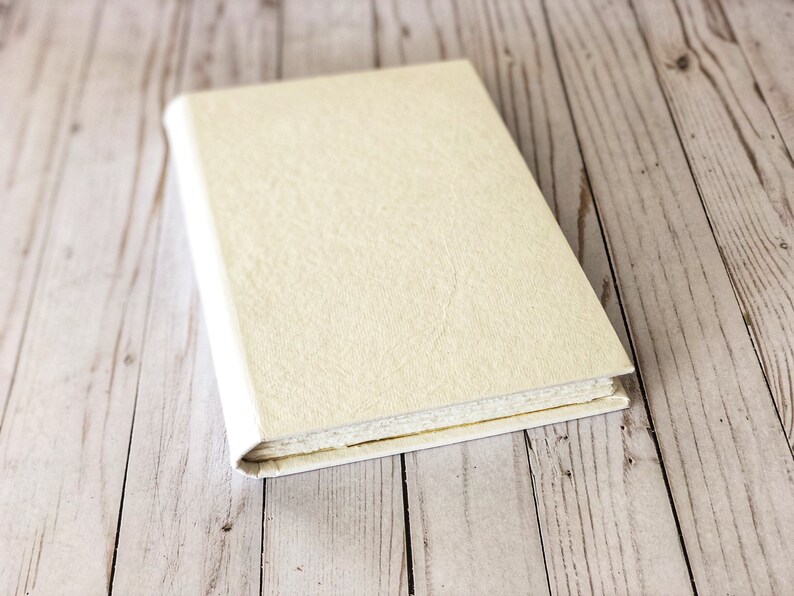 May include: A closed, off-white journal with a textured cover sits on a light wood surface.