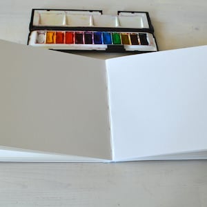 Puede incluir: Un cuaderno de bocetos abierto con p&aacute;ginas blancas en blanco. El cuaderno de bocetos est&aacute; cerrado en el lado izquierdo y abierto en el lado derecho, revelando las p&aacute;ginas en blanco. El cuaderno de bocetos est&aacute; sobre una superficie blanca.