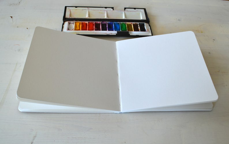 Puede incluir: Un cuaderno de bocetos abierto con p&aacute;ginas blancas en blanco. El cuaderno de bocetos est&aacute; cerrado en el lado izquierdo y abierto en el lado derecho, revelando las p&aacute;ginas en blanco. El cuaderno de bocetos est&aacute; sobre una superficie blanca.