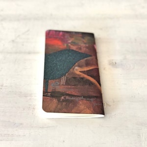 May include: A small, brown notebook with a colorful abstract design on the cover. The design features a dark blue shape, a light brown shape, and a red and orange shape. The text "everything is possible" is written on the cover.