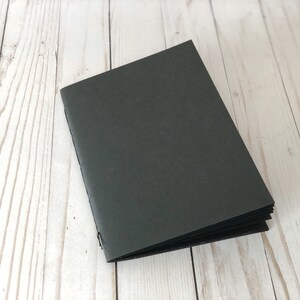 May include: A black notebook with a textured cover. The notebook is closed and lying on a white wooden surface.