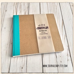 Puede incluir: Un diario marr&oacute;n con una encuadernaci&oacute;n turquesa y una etiqueta blanca con el texto "To LIVE a Creative LiFe WE MUST LOSE the fear of being WRONG THE JOURNAL SHOP" y la direcci&oacute;n del sitio web "WWW.JOURNALSHOP.ETSY.COM".