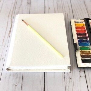 May include: A white blank sketchbook with a yellow paintbrush lying on top of it. A watercolor paint set with a variety of colors is to the right of the sketchbook.
