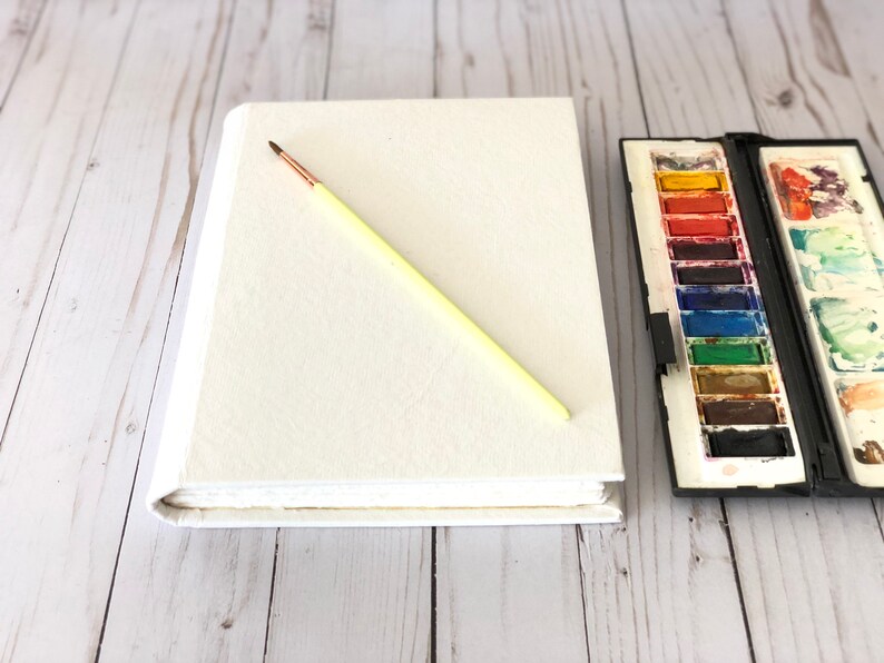 May include: A white blank sketchbook with a yellow paintbrush lying on top of it. A watercolor paint set with a variety of colors is to the right of the sketchbook.