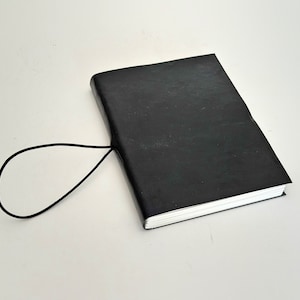 May include: A black leather-bound journal with a black elastic closure. The journal is closed and lying on a white surface.