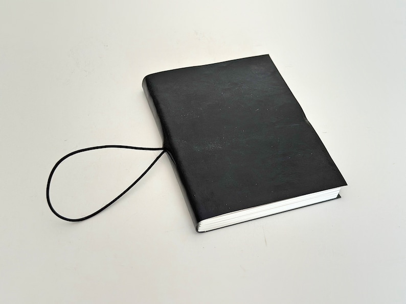 May include: A black leather-bound journal with a black elastic closure. The journal is closed and lying on a white surface.