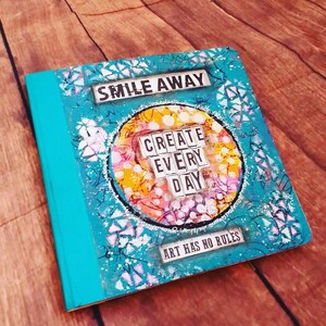 Puede incluir: Una libreta turquesa con un dise&ntilde;o colorido y el texto "Smile Away" y "Create Every Day" en la portada. El texto "Art Has No Rules" tambi&eacute;n est&aacute; en la portada.