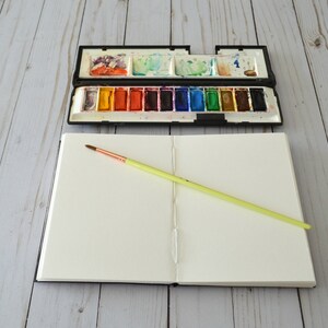 May include: An open sketchbook with a blank page and a yellow paintbrush lying on top. A closed watercolour paint palette with a variety of colours is visible in the background.