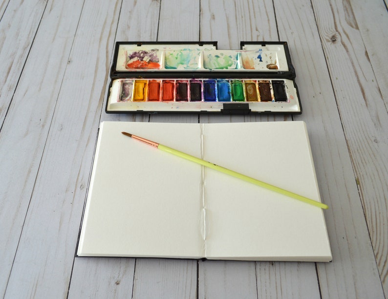 May include: An open sketchbook with a blank page and a yellow paintbrush lying on top. A closed watercolour paint palette with a variety of colours is visible in the background.