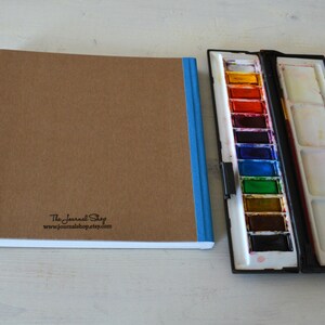Puede incluir: Un cuaderno de bocetos marr&oacute;n con una encuadernaci&oacute;n azul y el texto "The Journal Shop www.journal-shop.com" impreso en la portada. Un juego de pintura de acuarela negro con una fila de 12 bandejas de pintura de colores est&aacute; al lado del cuaderno de bocetos.