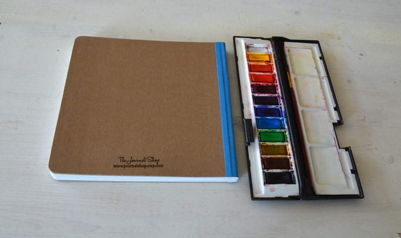 Puede incluir: Un cuaderno de bocetos marr&oacute;n con una encuadernaci&oacute;n azul y el texto "The Journal Shop www.journal-shop.com" impreso en la portada. Un juego de pintura de acuarela negro con una fila de 12 bandejas de pintura de colores est&aacute; al lado del cuaderno de bocetos.