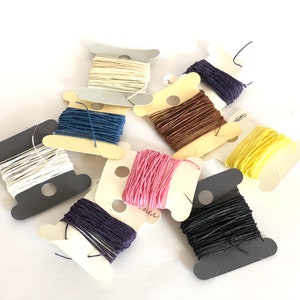 May include: Assortment of eight spools of thread in various colors, including white, black, navy blue, brown, yellow, pink, and purple. The spools are arranged on a white surface.