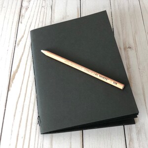 May include: A black notebook with a wooden pencil on top. The pencil is labeled "Derwent Charcoal White 558".