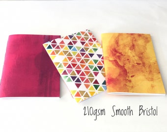 May include: Three notebooks with different covers. The first notebook has a solid pink cover. The second notebook has a white cover with a colorful geometric pattern of triangles. The third notebook has a yellow cover with a watercolor-style design in shades of orange and red. The text "210gsm Smooth Bristol" is written below the notebooks.