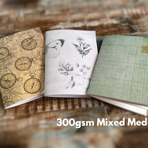May include: Three notebooks with 300gsm mixed media paper. The left notebook features a vintage compass map pattern. The center notebook displays black and white botanical illustrations of a butterfly and plants. The right notebook has a green grid pattern.