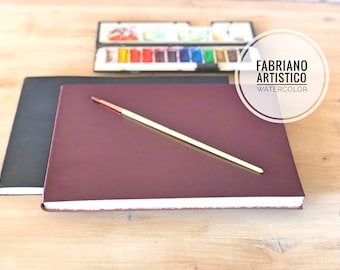 May include: A burgundy watercolor sketchbook with a white edge and a paintbrush lying on top. The sketchbook is labeled "Fabriano Artistico Watercolor".