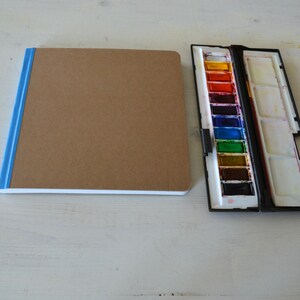 Puede incluir: Un cuaderno de bocetos marr&oacute;n con una encuadernaci&oacute;n azul y un juego de pintura de acuarela con una caja negra. El juego de pintura tiene 12 pastillas de acuarela y un pincel.
