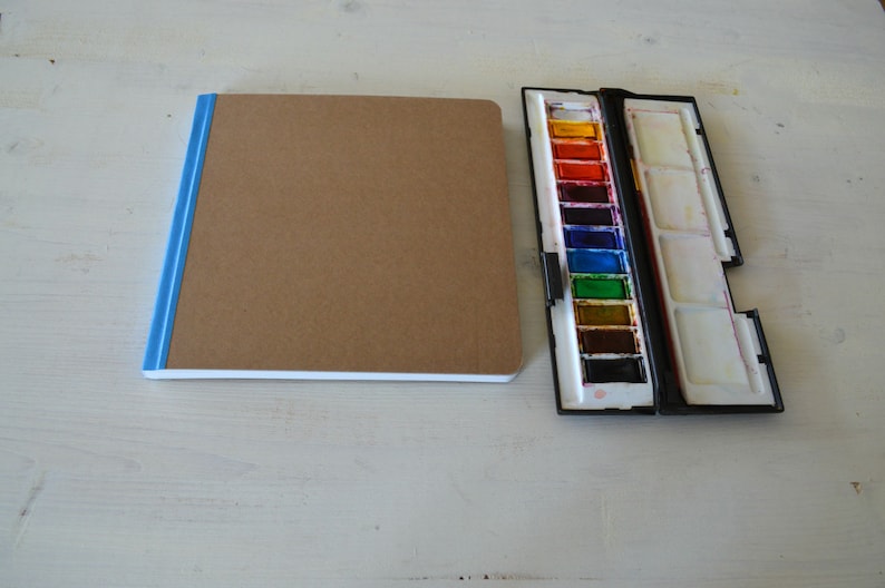 Puede incluir: Un cuaderno de bocetos marr&oacute;n con una encuadernaci&oacute;n azul y un juego de pintura de acuarela con una caja negra. El juego de pintura tiene 12 pastillas de acuarela y un pincel.