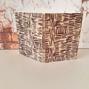 Puede incluir: Un pequeño libro hecho a mano con una cubierta de patrones marrones y blancos. La cubierta está hecha de papel y tiene una superficie texturizada. El libro está encuadernado con alambre de cobre.