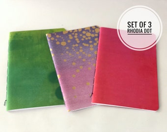 May include: Set of three Rhodia dot notebooks. The notebooks have different colored covers: green, purple with gold dots, and red.