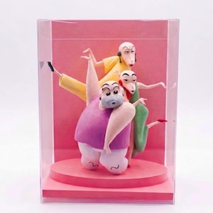 Op de afbeelding: Een doorzichtige plastic vitrine met drie gestileerde kleifiguren op een ronde roze voet. De figuren omvatten een groot persoon in een paars hemd en witte broek, een persoon in een gele outfit en een persoon in een groene jurk, allen in een pose.