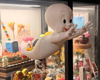 Può includere: Una statuetta bianca di Casper il fantasma amichevole con una finitura opaca liscia, grandi occhi neri e una bocca sorridente, raffigurata in una posa di volo con le braccia tese in avanti, posizionata davanti a una vetrina piena di vari giocattoli.