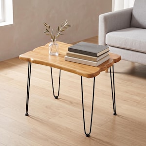 May include: A rectangular wooden side table with a natural live edge and four thin black metal hairpin legs. A clear glass vase containing a small green leafy branch and a stack of three books with gray and beige covers sit on the tabletop.