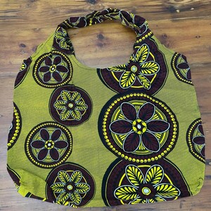 May include: A hobo-style shoulder bag featuring a vibrant mustard yellow background with a repeating pattern of large circular motifs. Each circle contains a dark brown and yellow six-petaled flower design with a central white dot accent.