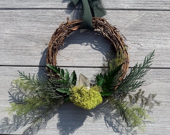 May include: A circular wreath made of brown twisted twigs, adorned with green fern fronds, evergreen sprigs, and a bright lime green moss cluster. A clear, pointed quartz crystal is nestled within the moss, and a dark green fabric ribbon is tied at the top.