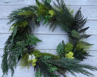 May include: A circular wreath constructed from dark green fern fronds, evergreen cedar sprigs, and patches of bright lime green preserved moss. The arrangement features a mix of textures with deep forest green foliage and light green moss clusters.