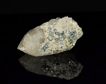 May include: A clear, pointed quartz crystal specimen featuring a rough, textured base embedded with numerous small, metallic, dark gray mineral inclusions. The crystal displays a translucent, jagged structure with a natural, unpolished surface.
