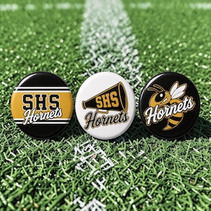 May include: Three circular pin-back buttons featuring SHS Hornets branding. The left button is black with gold stripes and text. The center white button displays a megaphone graphic with text. The right black button features a hornet illustration.