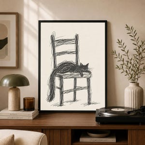 May include: A framed wall art print featuring a black ink scribble drawing of a cat resting on a wooden chair. The artwork is rendered in a loose, gestural style with dark lines against a light cream background, enclosed in a simple black frame.