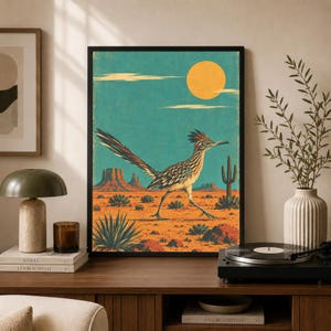 Peut inclure: Une affiche encadrée représentant un grand géocoucou marchant dans un paysage désertique avec des mesas, des cactus et un grand soleil jaune sur un ciel sarcelle. L'illustration utilise une palette de couleurs chaudes orange, brun et jaune.