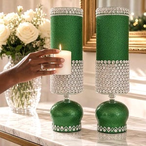 Peut inclure: Deux bougeoirs cylindriques avec une finition à paillettes vert vif, ornés de bandes de strass transparents et étincelants sur le haut, le milieu et la base. Chaque bougeoir repose sur un socle arrondi recouvert de paillettes vertes.