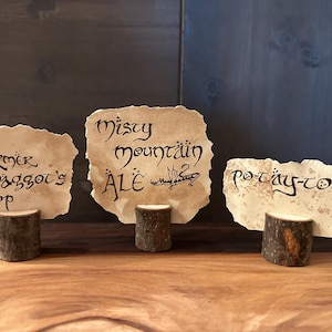 Puede incluir: Tres piezas rectangulares de papel pergamino envejecido de color blanco roto con bordes irregulares y rasgados, cada una sostenida por una pequeña base de tronco de madera cilíndrica. El papel presenta caligrafía negra con los textos Farmer Maggot's Crop, Misty Mountain Ale y Po-tay-toes.
