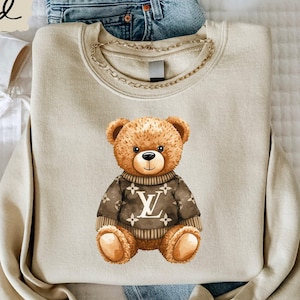 May include: A sand-colored crewneck sweatshirt featuring a graphic of a brown teddy bear wearing a dark brown sweater with a repeating white floral pattern and a central white logo. The sweatshirt is displayed with blue jeans and white sneakers.