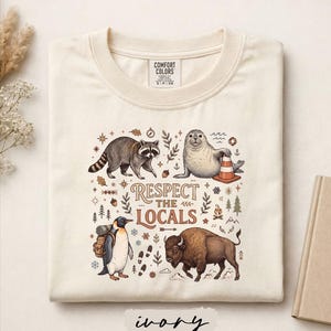 May include: An ivory t-shirt featuring a graphic print with a raccoon, a seal with a traffic cone, a penguin with a backpack, and a bison. The text "RESPECT THE LOCALS" is centered among small illustrations of trees, leaves, and a campfire.