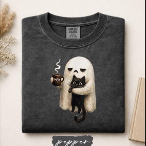 May include: A charcoal gray crewneck t-shirt featuring a graphic of a white ghost holding a brown mug with the text MEH and a black cat. The shirt has a Comfort Colors label inside the collar and a folded, worn-in fabric texture throughout.
