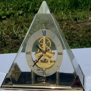 May include: A pyramid-shaped Seiko Quartz mantel clock with a clear acrylic casing and a gold-tone metal base. The clock face features a white circular dial with black Roman numerals, gold-tone hands, and a visible internal gear mechanism.